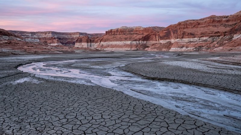 A drained reservoir bed of cracked mud with a pale bathtub ring on the cliffs behind