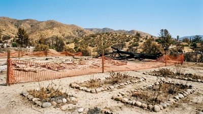 A cleared lot after a wildfire with orange safety fencing across bare soil and a charred fallen tree behind