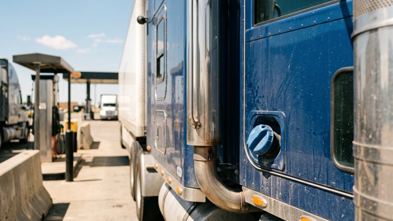 The chrome exhaust stack and blue diesel exhaust fluid cap on the side of a semi tractor at a truck stop.