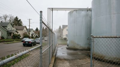 Two industrial storage tanks behind a chain-link fence with water mist drifting across them beside a residential street