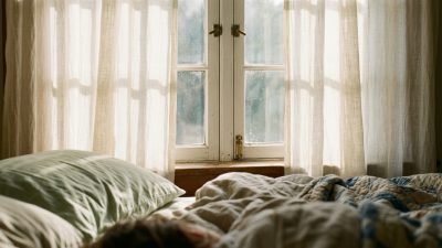 A closed bedroom window at night seen from inside, with streetlight bleeding orange through thin curtains and the edge of a pillow soft in the foreground.