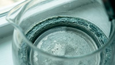 Looking down into a glass kettle at a thick teal and gray-green crust of limescale ringing the rim, with a pale chalky sheet of scale and darker grit across the base under a film of water.