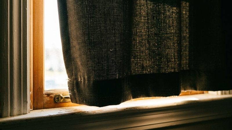 A heavy lined curtain drawn across an old wooden bedroom window, with morning light spilling around its edge and across the sill. That gap is the largest single light source in most bedrooms, and a lined curtain closes it in an afternoon.
