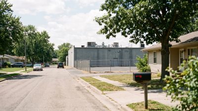 A long windowless gray building with rooftop cooling units behind a chain link fence at the end of a residential street.