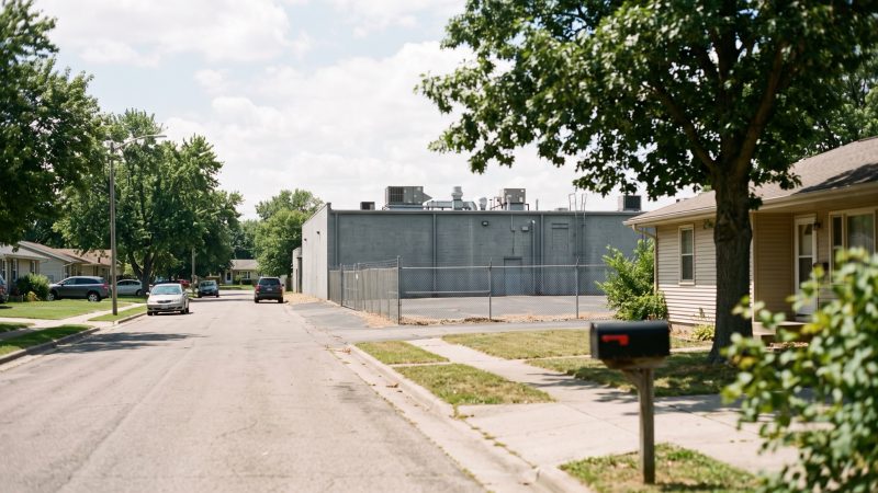A long windowless gray building with rooftop cooling units behind a chain link fence at the end of a residential street.