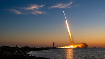 A white rocket climbing into a twilight sky on a bright exhaust plume, seen across coastal water