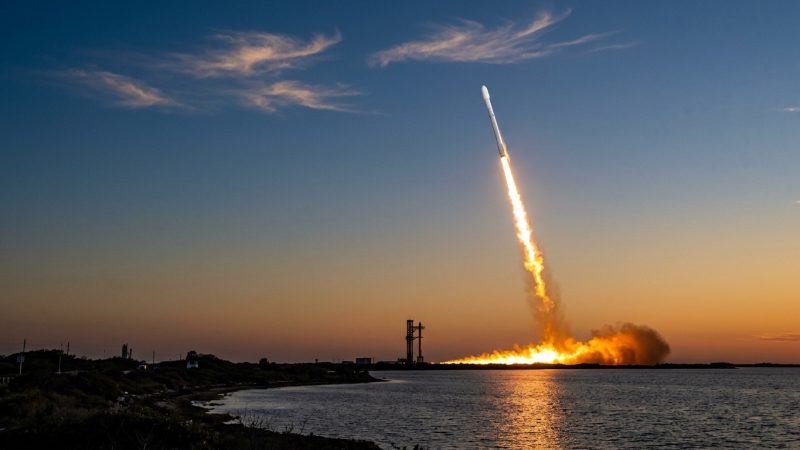 A white rocket climbing into a twilight sky on a bright exhaust plume, seen across coastal water