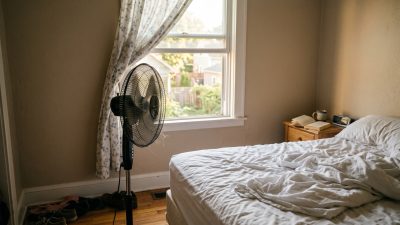 An oscillating floor fan turned toward a rumpled bed in a small bedroom at sunrise.