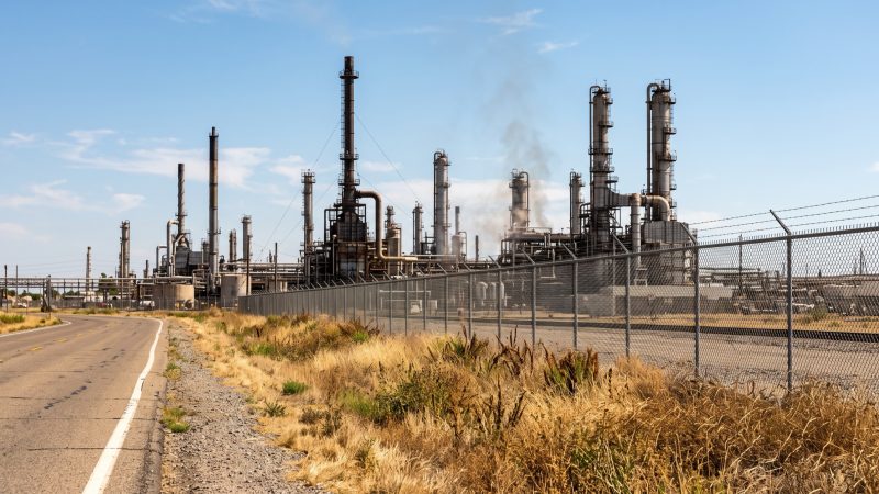 The stacks and pipework of an oil refinery behind a chain link fence, seen from a public road.