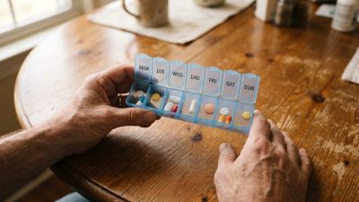 A pair of hands holding an open weekly pill organizer over a wooden table.