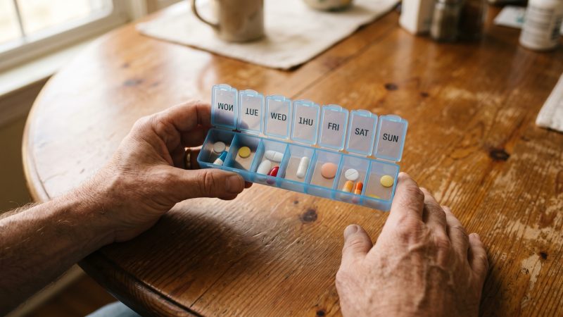 A pair of hands holding an open weekly pill organizer over a wooden table.